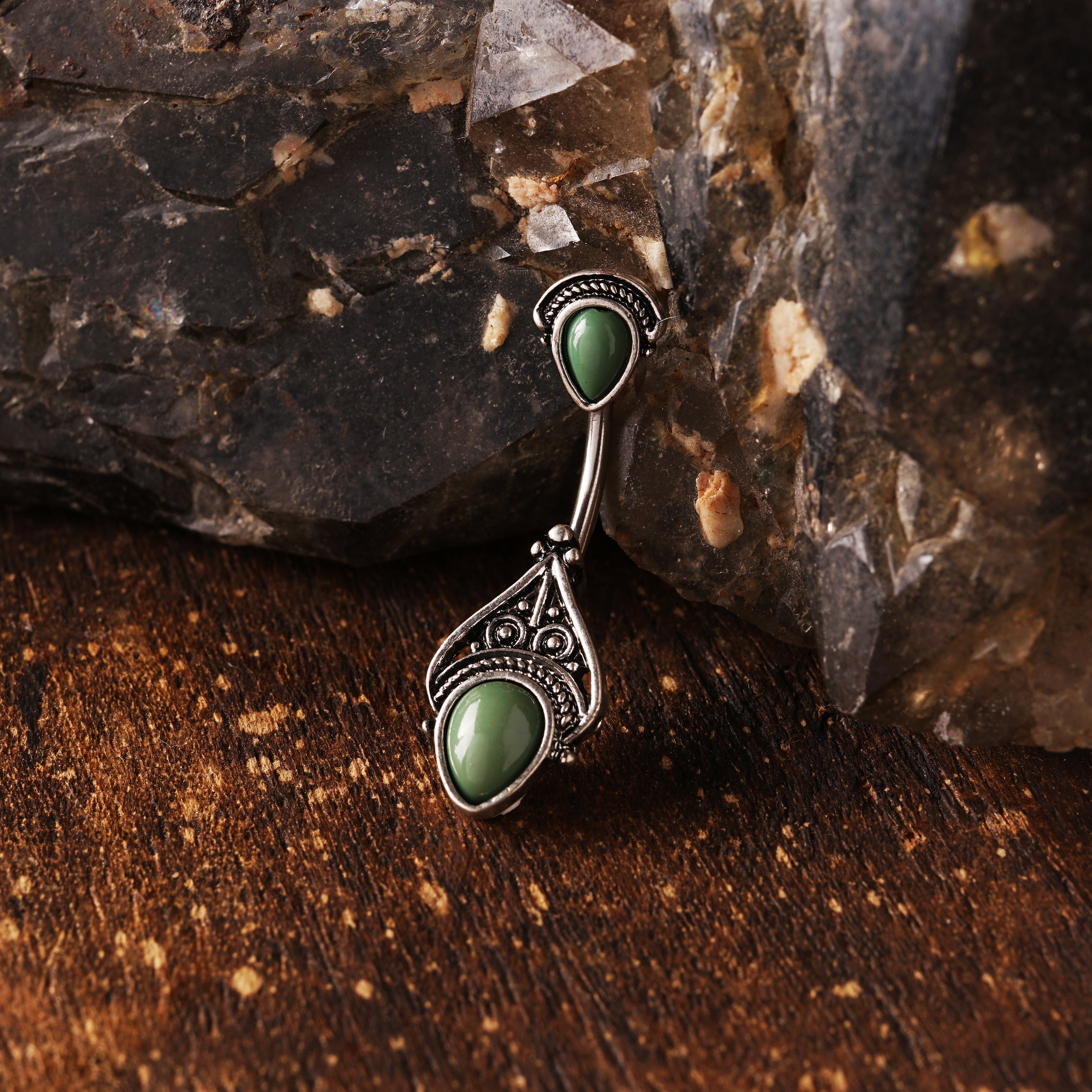 navel bar with silver toned goth ornament with two green stones one at the top and one at the bottom on a wooden background